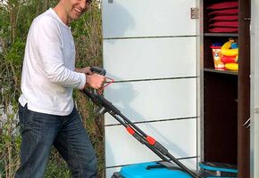 . Ein Mann schiebt einen Rasenmäher vor einen Schrank im Garten, der mit Gartenbedarf gefüllt ist.