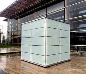 . Ein moderner, quadratischer Gartenschrank aus Glas und Metall steht vor einem Gebäude mit großen Fenstern.