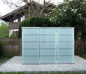 . Ein moderner Glas-Geräteschuppen vor einem Haus, umgeben von einem Garten mit Bäumen und Sträuchern.