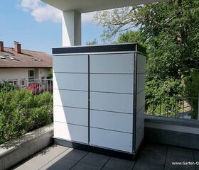 . Ein weißer Gartenschrank auf einer Terrasse, umgeben von Pflanzen und einem Haus im Hintergrund.