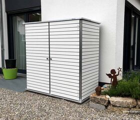 . Ein weißer Gartenschrank steht auf einer Kiesfläche neben einem Haus, mit Pflanzen und einer Engel-Deko daneben.