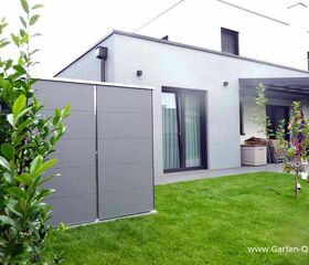 . Moderner Garten mit grauem Schrank, Terrasse, Pflanzen und gepflegtem Rasen vor einem Haus.