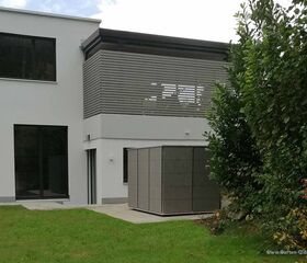 . Modernes Haus mit großen Fenstern, Terrasse und Gartenhütte, umgeben von grünem Rasen und Bäumen.