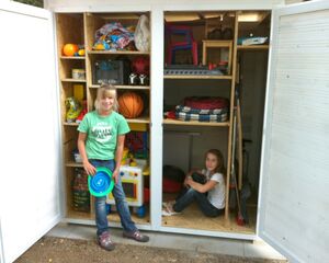 Ein Schuppen mit Spielzeug und Werkzeug. Zwei Kinder, eines steht mit Frisbee, das andere sitzt.