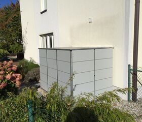 . Ein moderner, grauer Außenaufbewahrungsschrank steht an der Seite eines Hauses, umgeben von Gartenpflanzen.