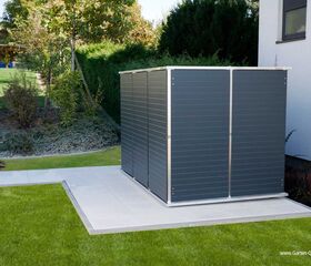 . Ein graues Gartenhaus steht neben einem Haus in einem gepflegten Garten mit Rasen und Bäumen.