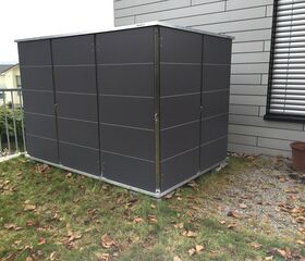 . Ein modernes, graues Gartenhaus steht auf Gras neben einem Gebäude mit grauen Wänden und einem Blumenkübel.