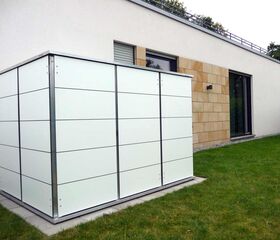 . Ein moderner weißer Gartenschuppen steht neben einem Haus auf einer Rasenfläche.