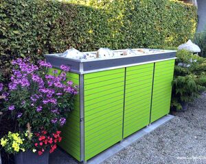 Ein grüner Mülleimer-Unterstand im Garten neben blühenden Pflanzen.