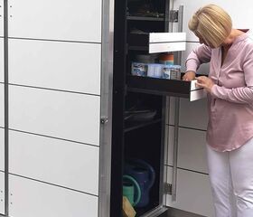 . Eine Person öffnet einen Gartenschrank mit Schubladen, Pflanzen und Gießkannen darin.