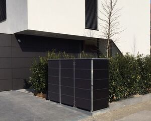 . Modernes Gebäude mit schwarzen Paneelen, umgeben von Pflanzen und einer Einfriedung.