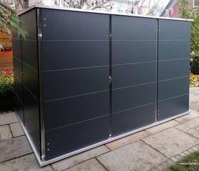 . Ein moderner Schuppen im Garten mit grauen Paneelen und umgeben von Pflanzen und Steinplatten.