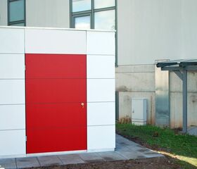 Ein rotes Tor in einer weißen Struktur vor einem Gebäude mit Fenstern, daneben eine Leiter.