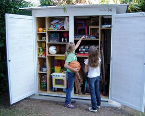 Ein geöffnetes Gartenhaus mit Spielzeugen und Werkzeugen. Zwei Kinder schauen hinein, eines hält einen Basketball.