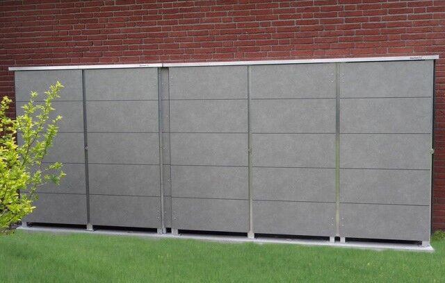 Ein moderner, grauer Lagerschrank steht vor einer Ziegelmauer auf einer grünen Rasenfläche mit einem kleinen Busch links.