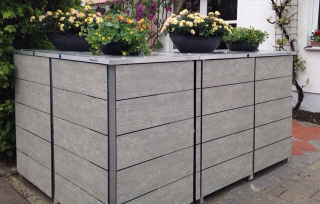 . Ein großer, moderner Mülleimer-Unterstand mit Holzoptik und Blumenkästen auf dem Dach.