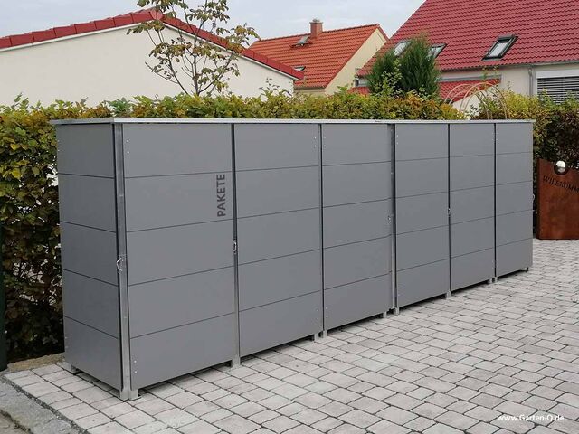 . Graue Paketkästen in einer gepflasterten Einfahrt, umgeben von Hecken und Häusern im Hintergrund.