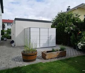 . Ein moderner Gartenschrank neben einem weißen Gebäude, umgeben von Pflanzen und Grünflächen.