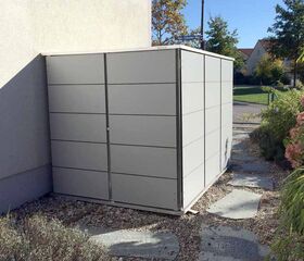 . Ein moderner, grauer Gartenschuppen steht seitlich an einem Haus, umgeben von Kies und Pflanzen.