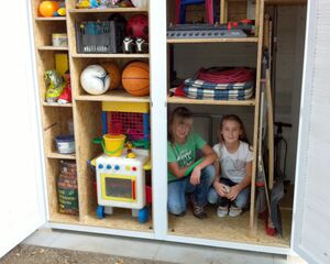 Ein Schuppen mit Spielsachen, Bällen, Stühlen und zwei Kindern, die im mittleren Fach sitzen.