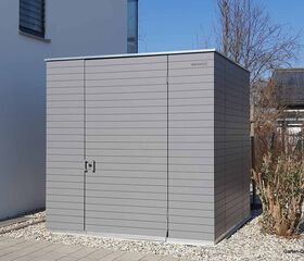 Graues Gartenhaus aus Holz vor einem modernen Gebäude.