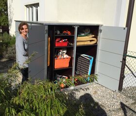 . Eine Person steht neben einem offenen Schrank im Freien mit Gartenartikeln und Taschen.