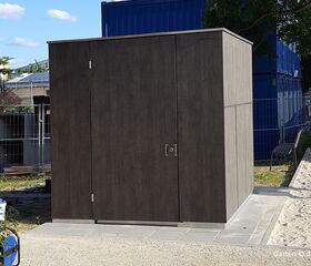 Ein moderner Gartenschuppen aus dunklem Holz auf einem Pflastersteinweg, umgeben von Bauzäunen und einer Bank.