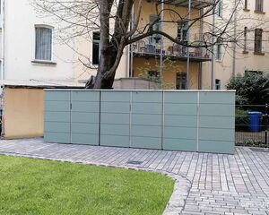 Ein Fahrradunterstand in einem Hof mit einem Baum, hinter einem Gebäude mit Balkonen.