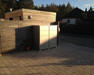 . Ein moderner Außenschrank steht auf einer gepflasterten Fläche neben einem Holzgebäude und einer kleinen Pflanze.