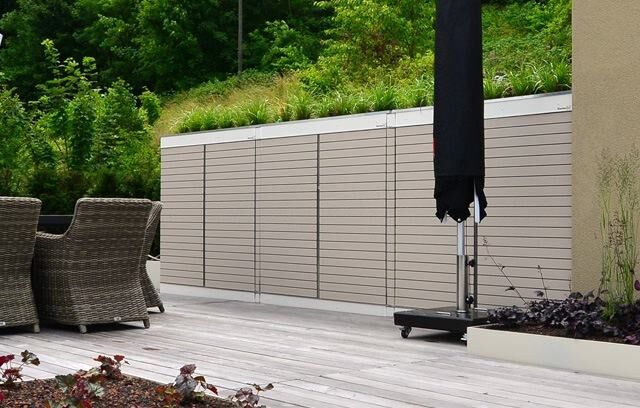 . Gartenmöbel auf Terrasse mit Holzfußboden, dahinter Sichtschutzzaun, schwarzer Sonnenschirm, umgeben von Pflanzen.