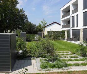 . Ein moderner Garten mit Rasen, Pflastersteinen, Grünpflanzen, einem Gartenschuppen und einer Terrasse vor einem Gebäude.