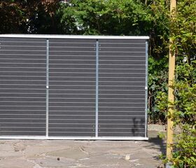 . Ein Solarmodul steht im Garten, umgeben von Pflanzen und einem Holzpfosten.