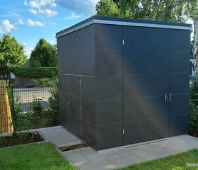 Ein moderner, dunkelgrauer Gartenschuppen steht auf gepflastertem Boden, umgeben von Grün und einem Zaun.