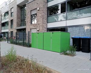 Wohngebäude mit Glasfront, Fahrrädern und grünem Schrank auf dem Gehweg.