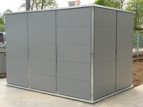 Eine graue Metallhütte steht auf einem gepflasterten Platz im Freien, umgeben von Bäumen und Zaun.