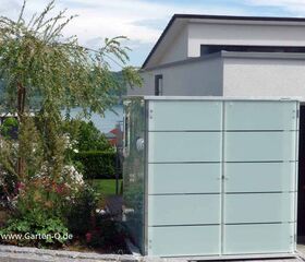 . Ein moderner Gartenschrank aus Glas mit Pflanze daneben, vor einem Haus mit Seeblick.
