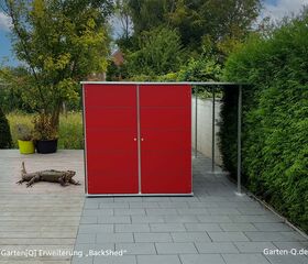 Rote Gartenhütte „BackShed“ mit einem großen Leguan davor, umgeben von Pflanzen.