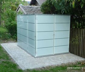 . Ein moderner Gartenhaus mit Glaswänden steht auf einem gepflasterten Bereich neben Bäumen und einem Holzzaun.