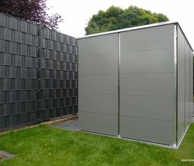 . Ein graues Gartengerätehaus steht auf einem Rasen neben einem schwarzen Zaun.