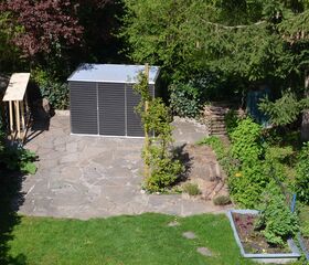 . Ein Garten mit Pflastersteinen, einem Schuppen, einem Komposthaufen und verschiedenen Pflanzen.