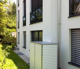 . Weiße Gartenbox neben einem modernen Gebäude mit abgedunkelten Fenstern, umgeben von einem gepflegten Rasen.