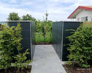 . Zwei moderne Gartenschuppen aus Metall, umgeben von grünen Büschen und einem gepflasterten Weg bei einem Haus.