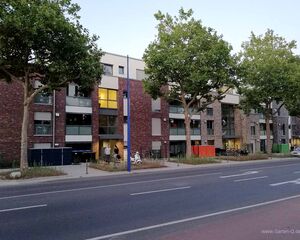 Mehrstöckiges Wohngebäude mit roter Klinkerfassade, Bäumen davor und Menschen auf dem Gehweg.