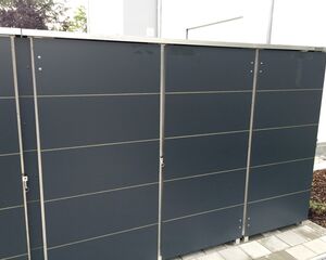 . Ein graues Metallmülltonnenverkleidung mit moderner Gestaltung, stehend auf Pflastersteinen, in einer Außenumgebung.