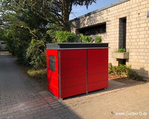 . Rote Paketbox neben Gebäude, bepflanzt auf dem Dach, steht auf gepflastertem Weg, umgeben von Bäumen und Sträuchern.