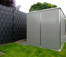 . Ein moderner, grauer Gartenschuppen neben einem Zaun, umgeben von grünem Rasen und Bäumen im Hintergrund.