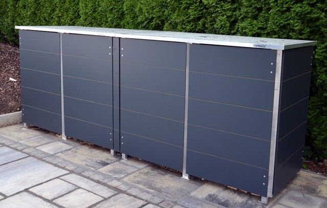 . Ein graues, modernes Mülltonnenhäuschen steht auf einem gepflasterten Boden vor einer Hecke.