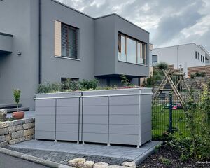 Moderne Häuser, graue Mülltonnenbox, Pflanzkübel oben, kleiner Garten mit Schaukeln und Bäumen.