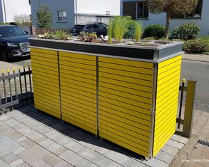 . Ein gelber Sichtschutz mit Bepflanzung oben, steht auf einem gepflasterten Bereich vor einem Wohngebäude.