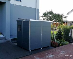 Ein moderner, grauer Außenlagerbehälter steht neben einem Gebäude, umgeben von Garten und Pflastersteinen.